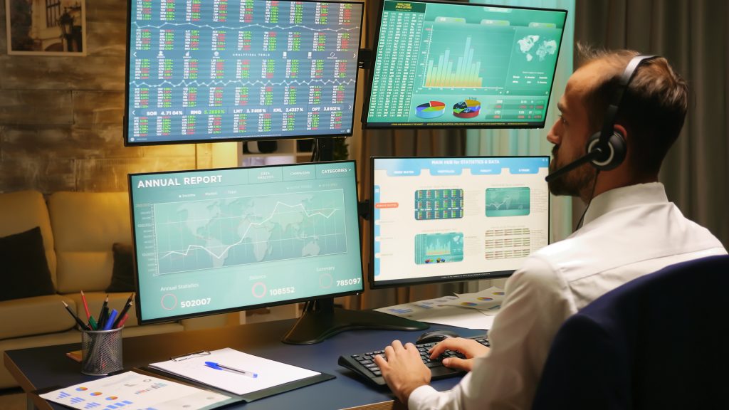 Back view of stock market trader looking at graphs on multiple monitors from home office.